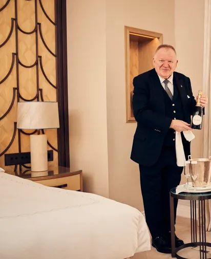 Butler standing in a well-appointed room, holding a bottle of champagne and dressed in formal uniform.