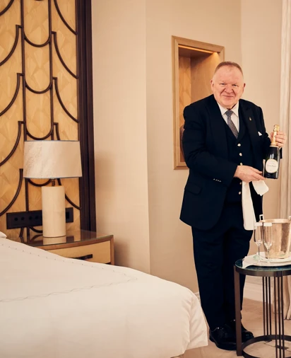 Butler standing in a well-appointed room, holding a bottle of champagne and dressed in formal uniform.