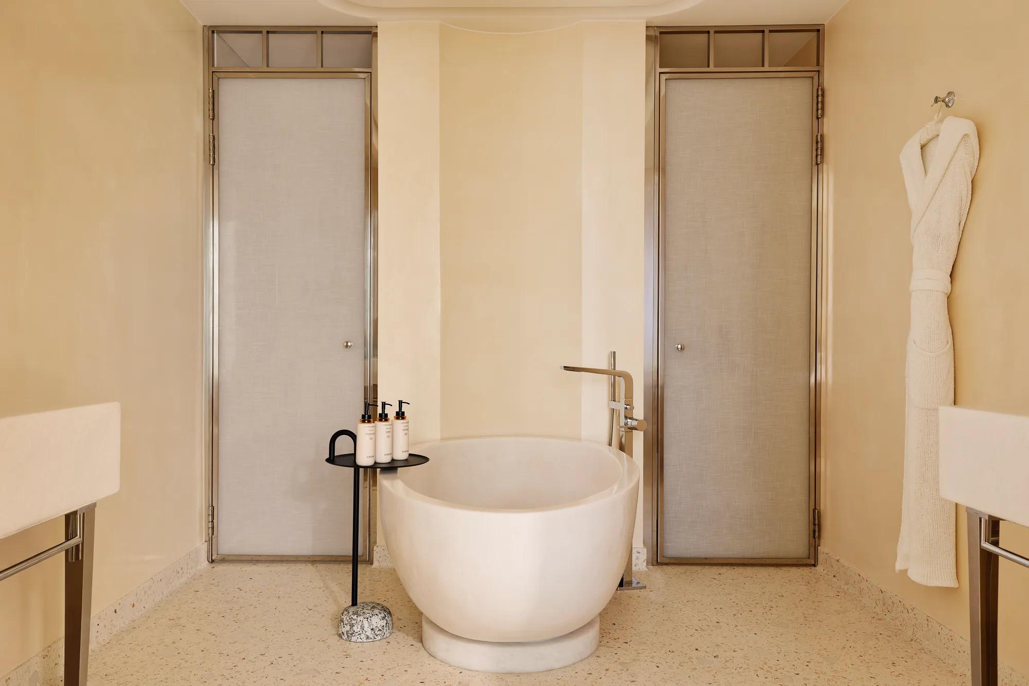 Luxurious bathroom with a sculpted freestanding bathtub, flanked by two doors, with a bathrobe hanging on the wall and amenities neatly placed on a side table.