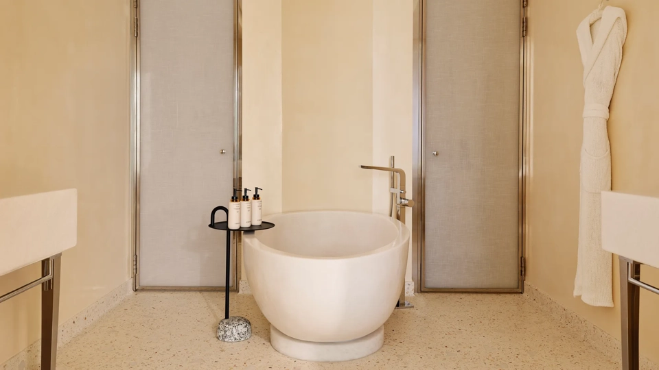 Luxurious bathroom with a sculpted freestanding bathtub, flanked by two doors, with a bathrobe hanging on the wall and amenities neatly placed on a side table.
