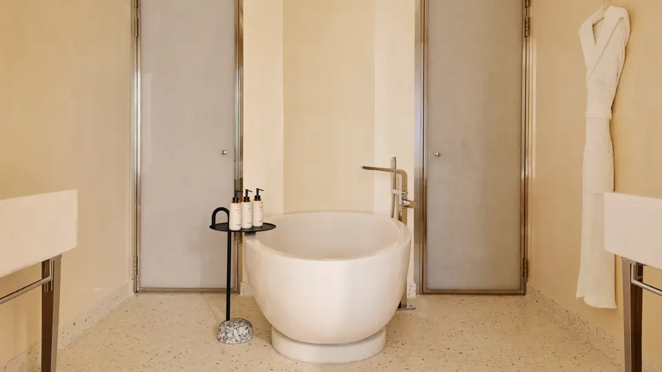 Luxurious bathroom with a sculpted freestanding bathtub, flanked by two doors, with a bathrobe hanging on the wall and amenities neatly placed on a side table.