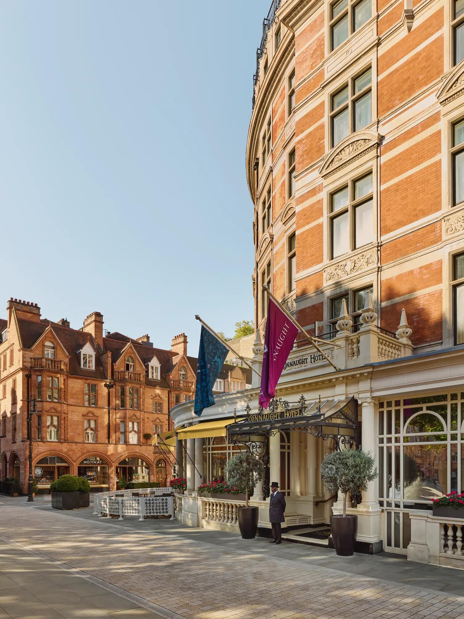 Exterior of a grand red-brick hotel with decorative stonework, entrance canopy and flags, set on a quiet paved street beside historic neighbouring buildings.
