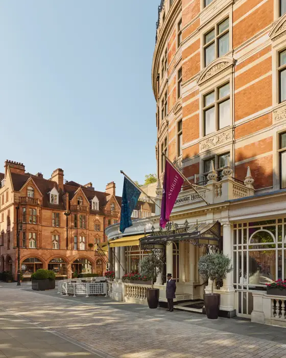 Exterior of a grand red-brick hotel with decorative stonework, entrance canopy and flags, set on a quiet paved street beside historic neighbouring buildings.