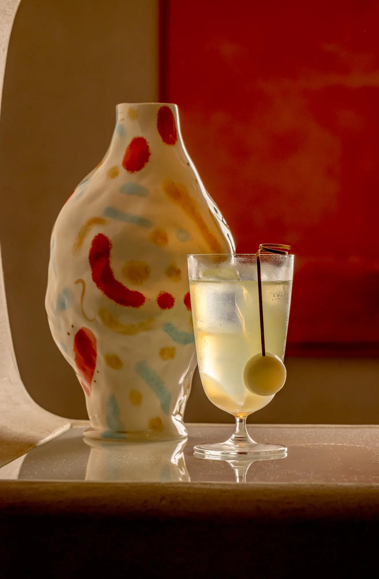 Stemmed cocktail with pale citrus drink and olive garnish beside a hand-painted ceramic vase on a softly lit bar.