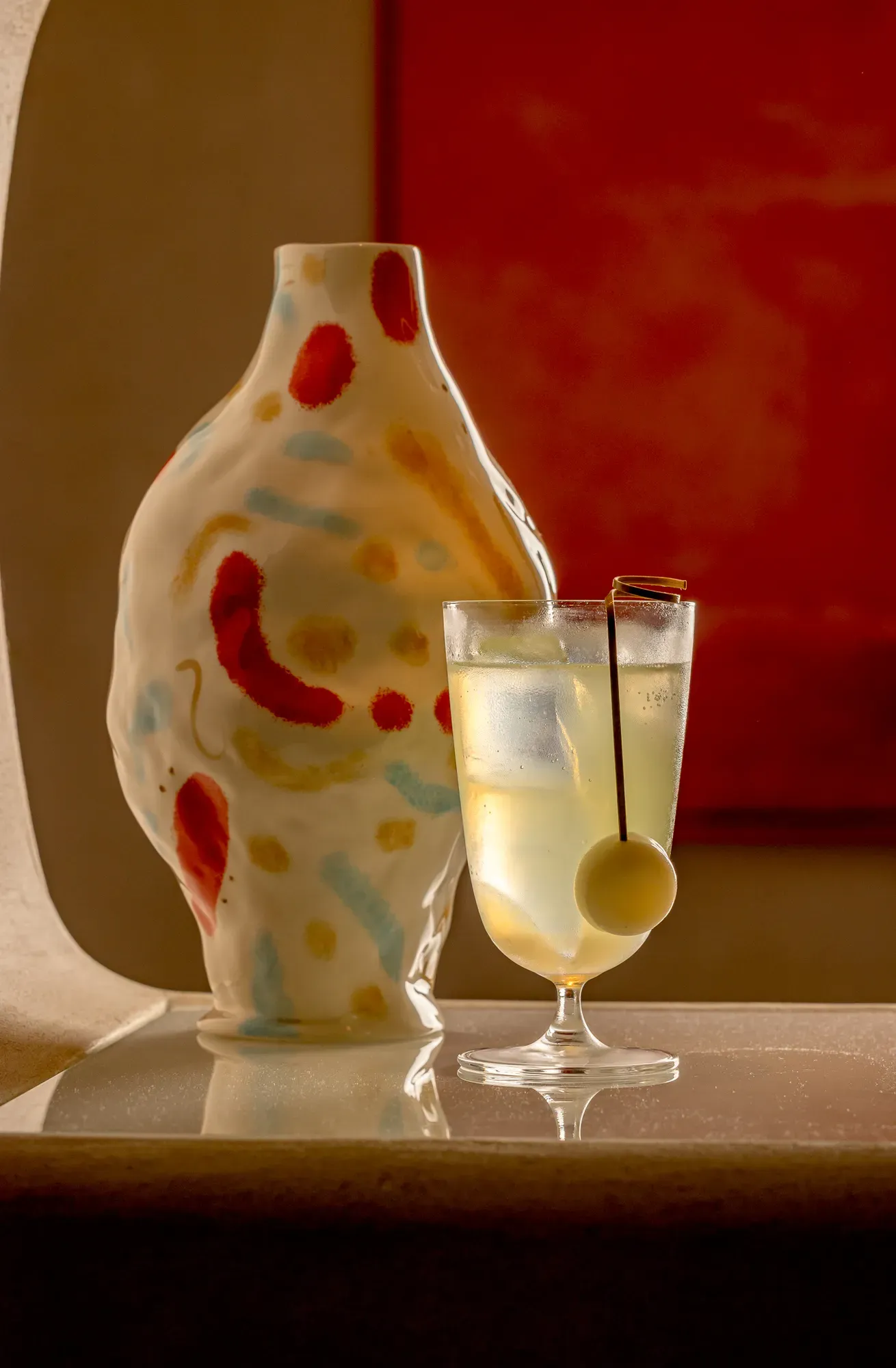 Stemmed cocktail with pale citrus drink and olive garnish beside a hand-painted ceramic vase on a softly lit bar.