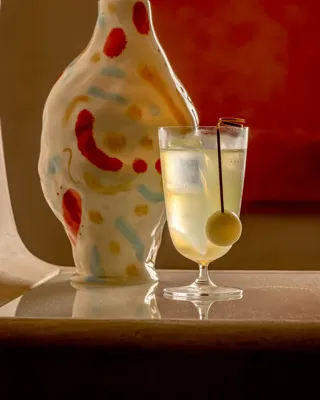 Stemmed cocktail with pale citrus drink and olive garnish beside a hand-painted ceramic vase on a softly lit bar.