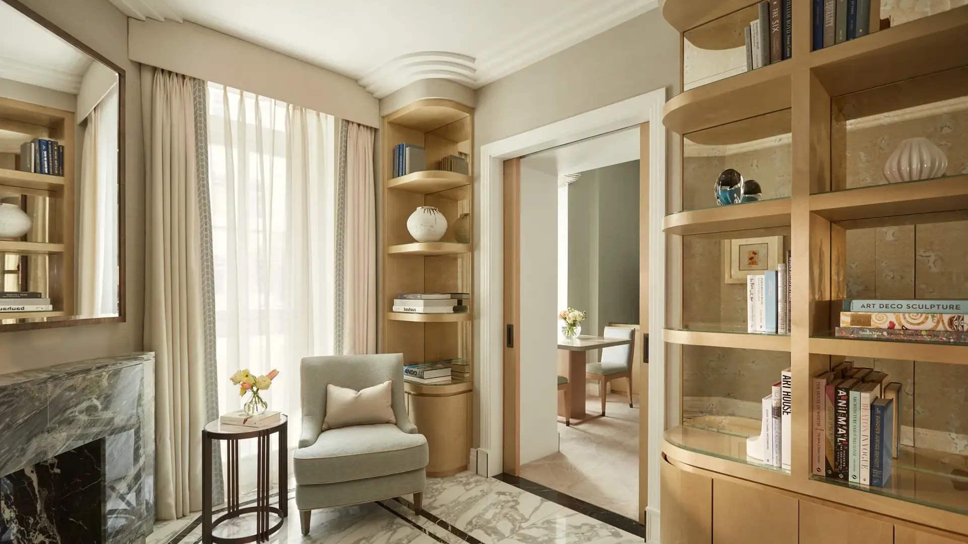 Elegant sitting area with marble flooring, a light grey armchair beside a marble fireplace, and built-in wooden bookshelves filled with books and decorative ceramics, leading to an adjoining room through sliding doors.