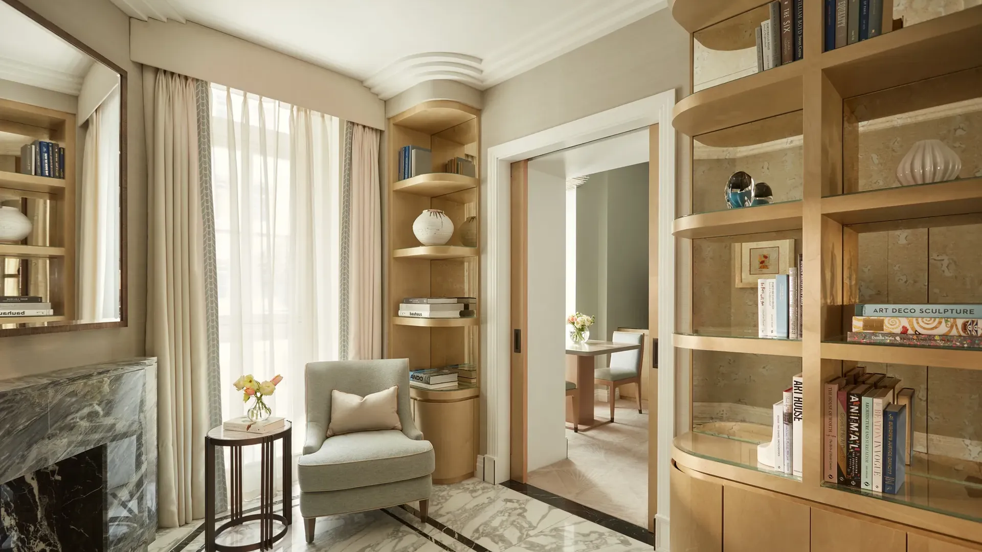 Elegant sitting area with marble flooring, a light grey armchair beside a marble fireplace, and built-in wooden bookshelves filled with books and decorative ceramics, leading to an adjoining room through sliding doors.