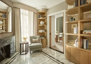 Elegant sitting area with marble flooring, a light grey armchair beside a marble fireplace, and built-in wooden bookshelves filled with books and decorative ceramics, leading to an adjoining room through sliding doors.
