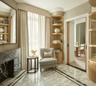 Elegant sitting area with marble flooring, a light grey armchair beside a marble fireplace, and built-in wooden bookshelves filled with books and decorative ceramics, leading to an adjoining room through sliding doors.