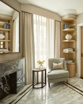 Elegant sitting area with marble flooring, a light grey armchair beside a marble fireplace, and built-in wooden bookshelves filled with books and decorative ceramics, leading to an adjoining room through sliding doors.