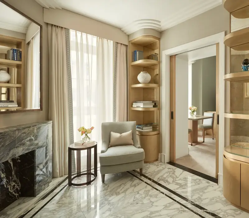 Elegant sitting area with marble flooring, a light grey armchair beside a marble fireplace, and built-in wooden bookshelves filled with books and decorative ceramics, leading to an adjoining room through sliding doors.