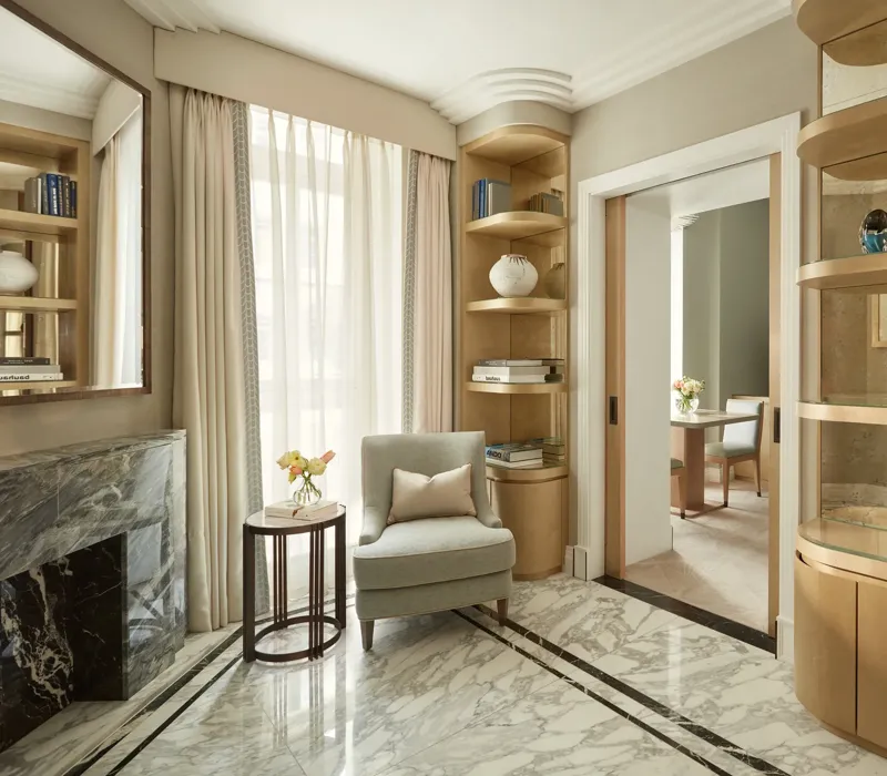 Elegant sitting area with marble flooring, a light grey armchair beside a marble fireplace, and built-in wooden bookshelves filled with books and decorative ceramics, leading to an adjoining room through sliding doors.