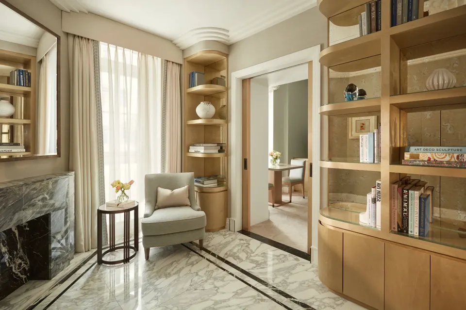Elegant sitting area with marble flooring, a light grey armchair beside a marble fireplace, and built-in wooden bookshelves filled with books and decorative ceramics, leading to an adjoining room through sliding doors.