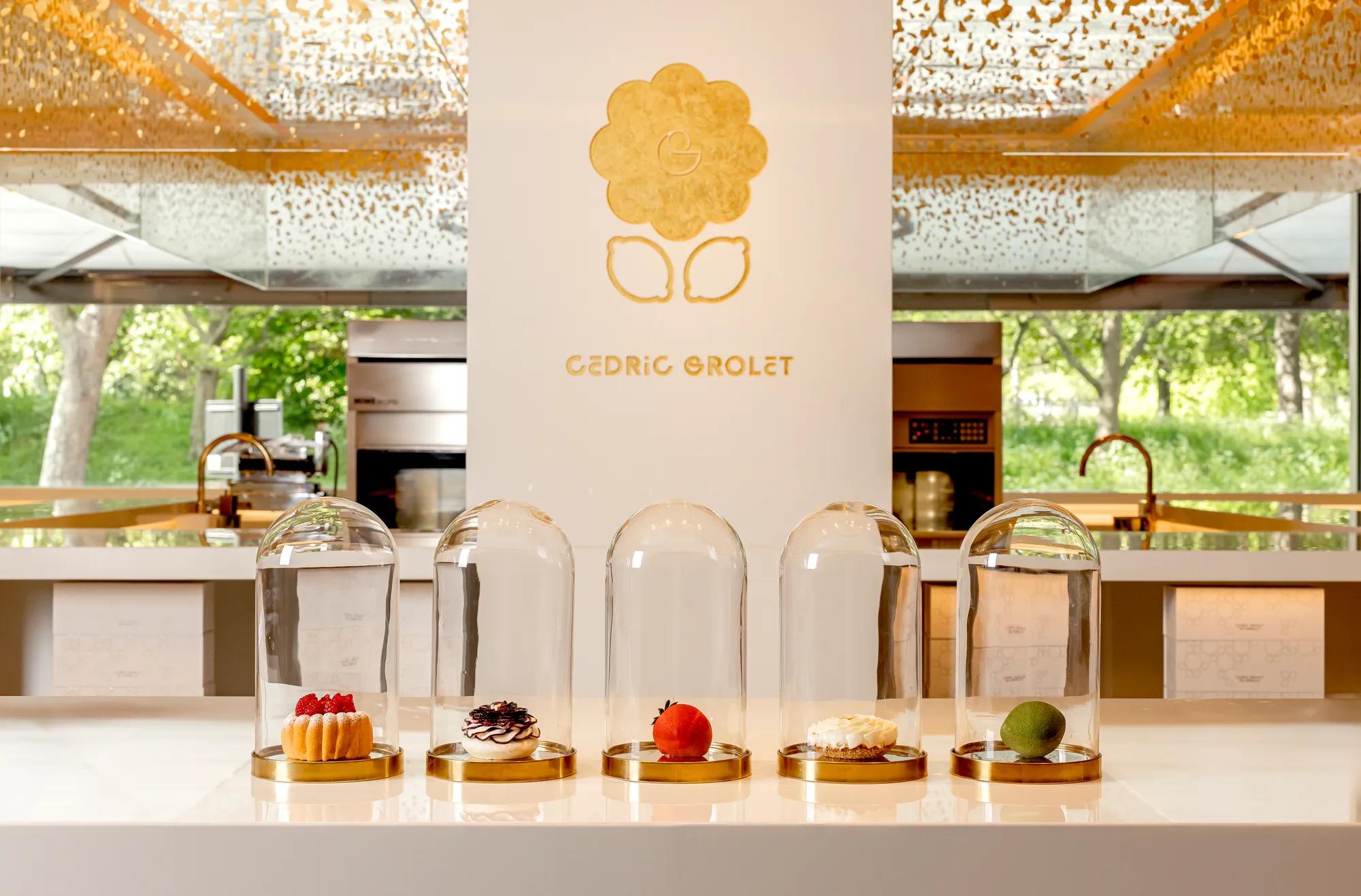 Five signature pastries, including fruit tarts and realistic fruit-shaped creations, displayed under glass domes on a white counter at Cédric Grolet, The Berkeley.