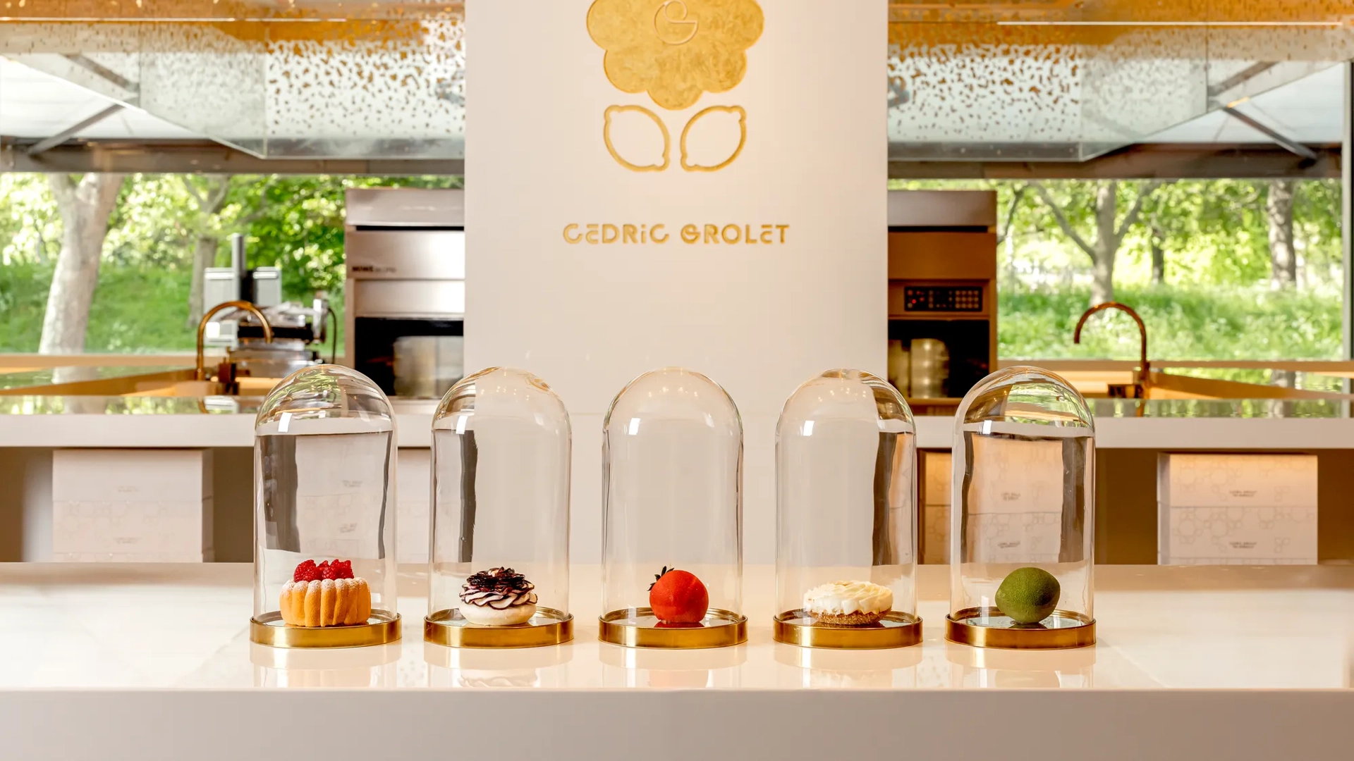 Five signature pastries, including fruit tarts and realistic fruit-shaped creations, displayed under glass domes on a white counter at Cédric Grolet, The Berkeley.