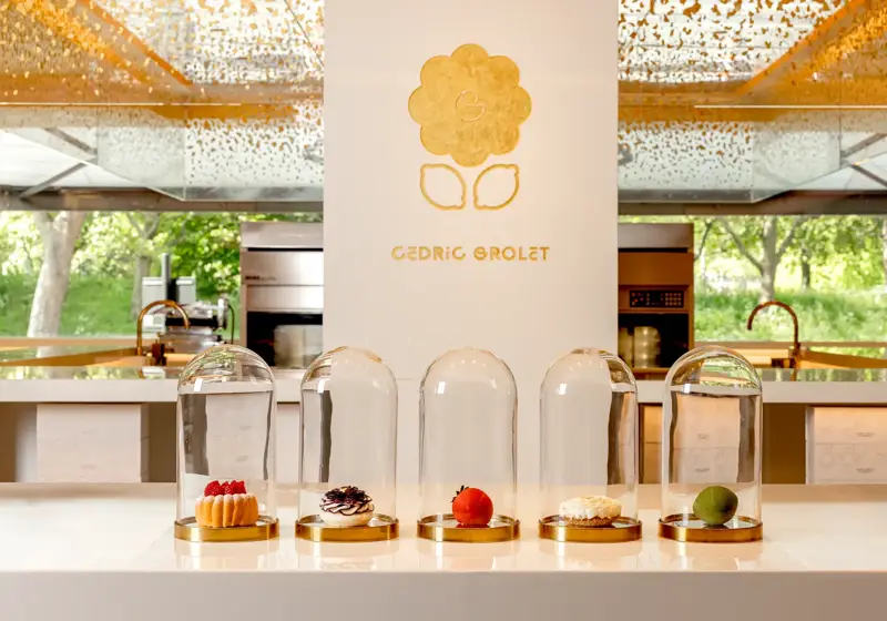 Five signature pastries, including fruit tarts and realistic fruit-shaped creations, displayed under glass domes on a white counter at Cédric Grolet, The Berkeley.