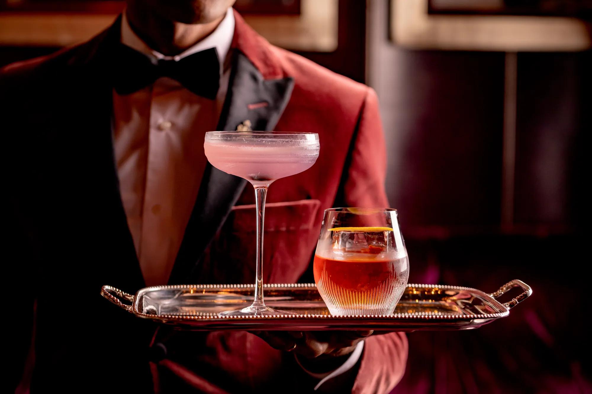 A bartender in a red velvet jacket and bow tie holding a silver tray with two elegant cocktails — a pale pink drink in a coupe glass and an amber drink in a tumbler — against a dimly lit bar background.