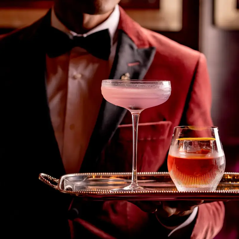 A bartender in a red velvet jacket and bow tie holding a silver tray with two elegant cocktails — a pale pink drink in a coupe glass and an amber drink in a tumbler — against a dimly lit bar background.