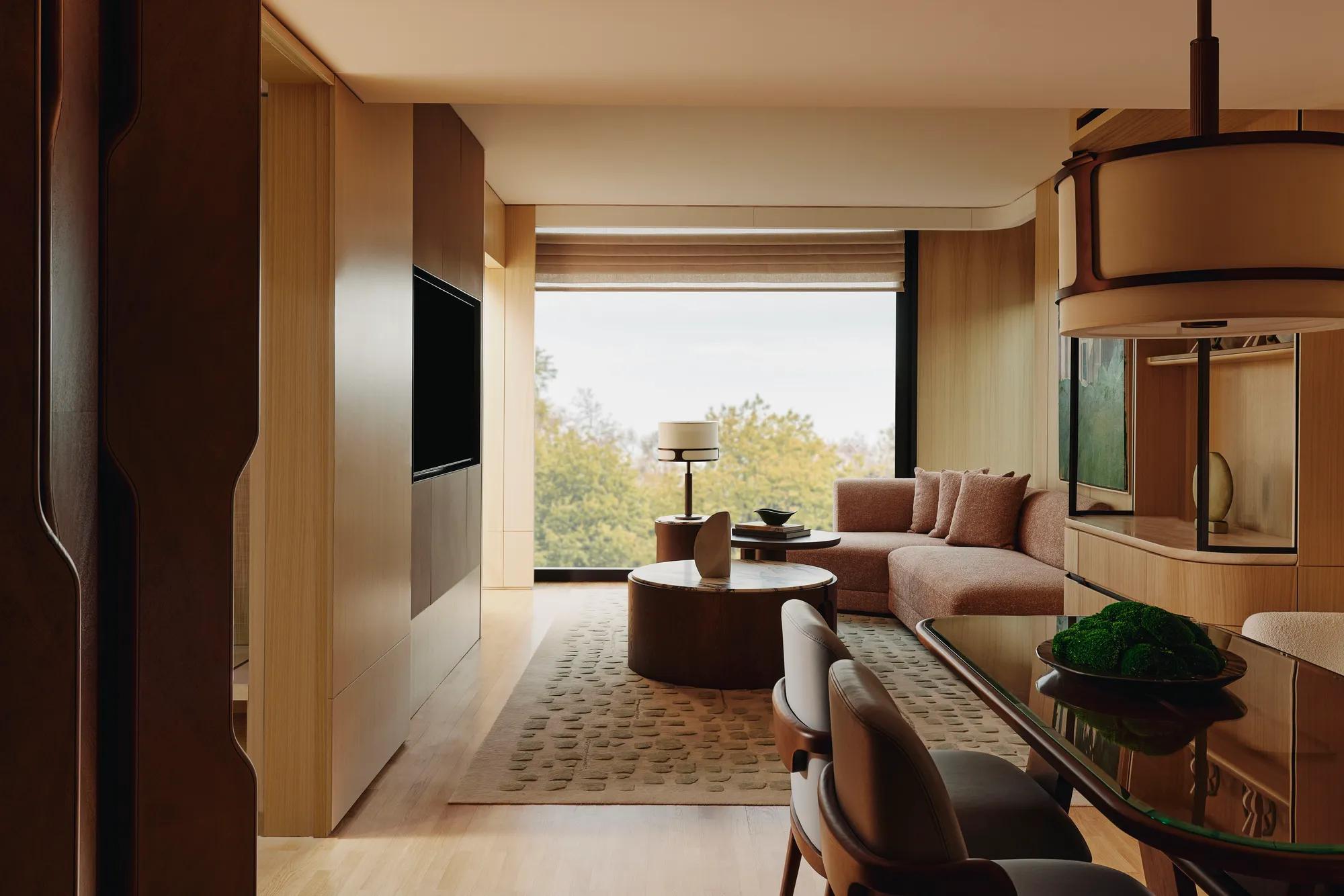 Spacious suite living and dining area with warm wood finishes, sofa seating, and large window overlooking trees.