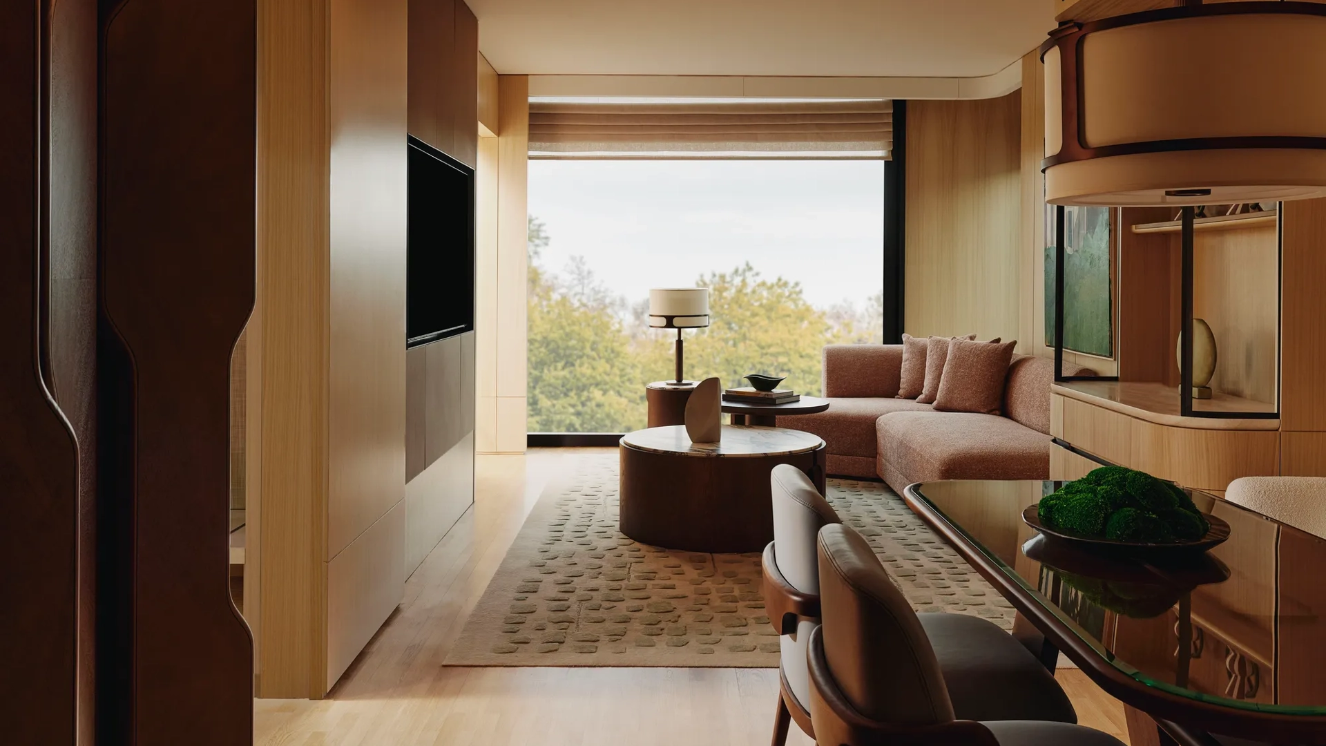 Spacious suite living and dining area with warm wood finishes, sofa seating, and large window overlooking trees.