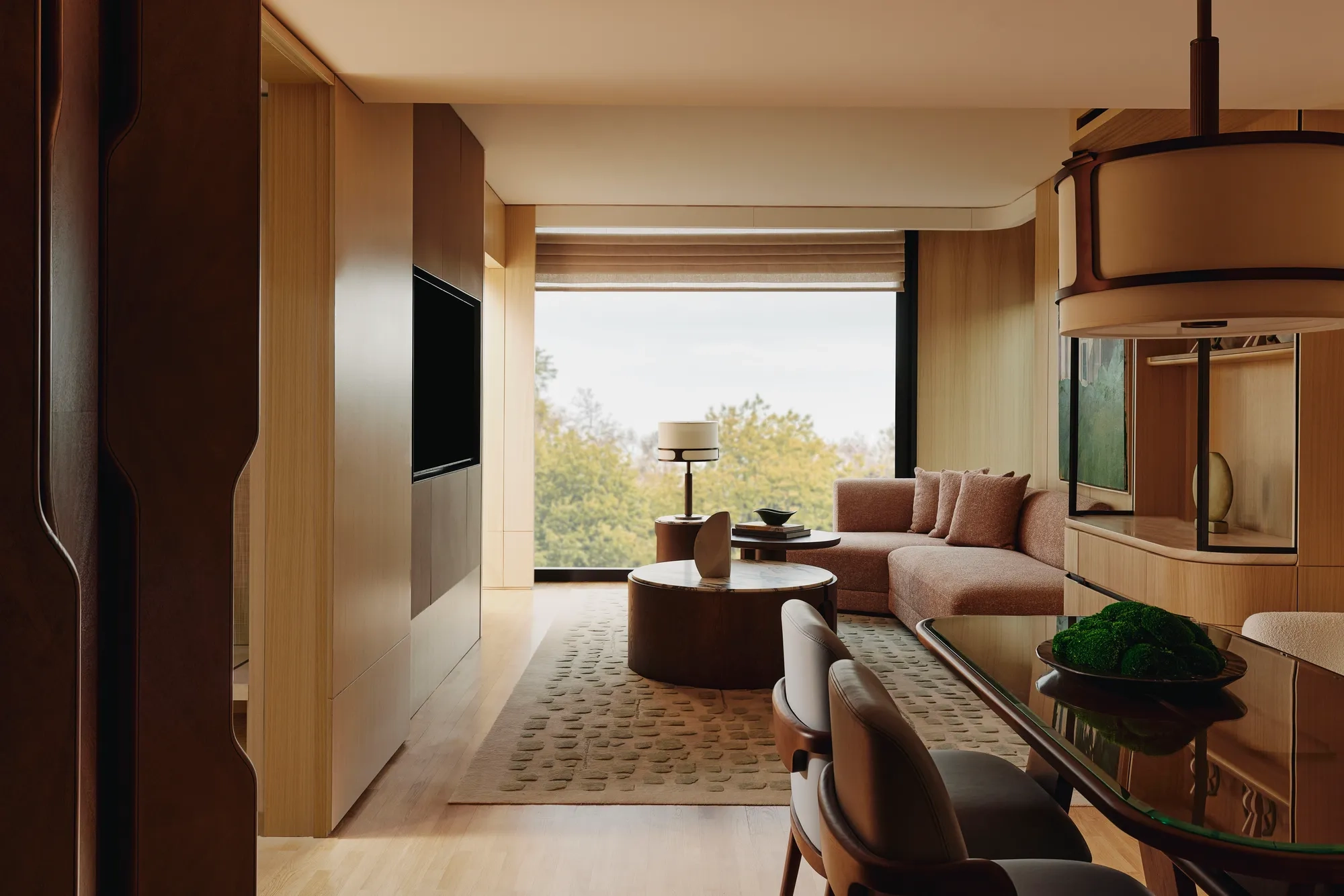 Spacious suite living and dining area with warm wood finishes, sofa seating, and large window overlooking trees.