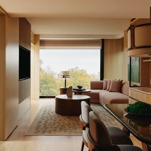 Spacious suite living and dining area with warm wood finishes, sofa seating, and large window overlooking trees.