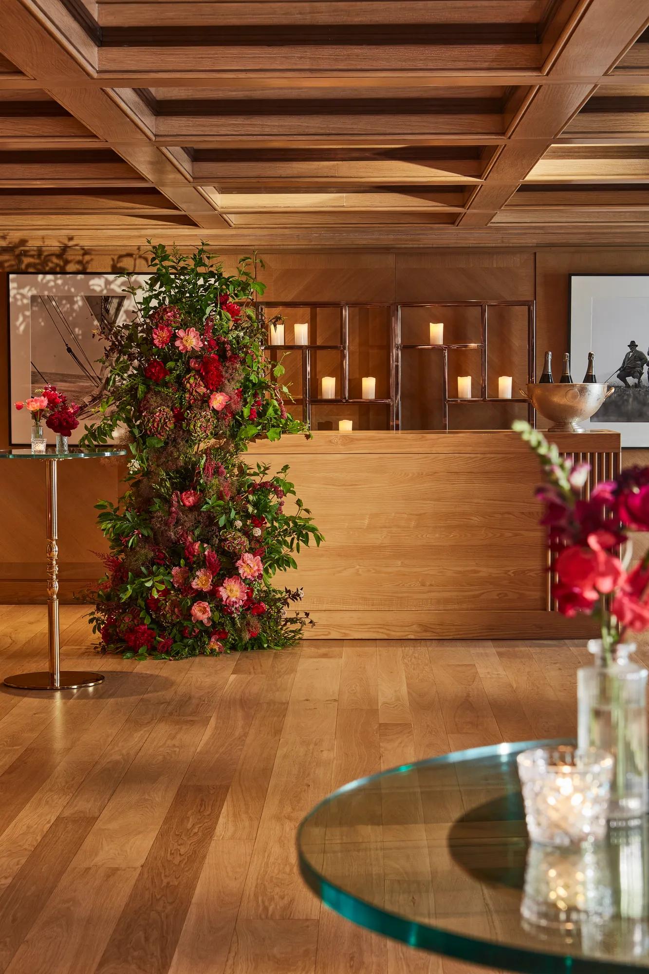 Wood-panelled reception space with a floral installation and candlelit display on a wooden counter, with small tables and flowers in the foreground.