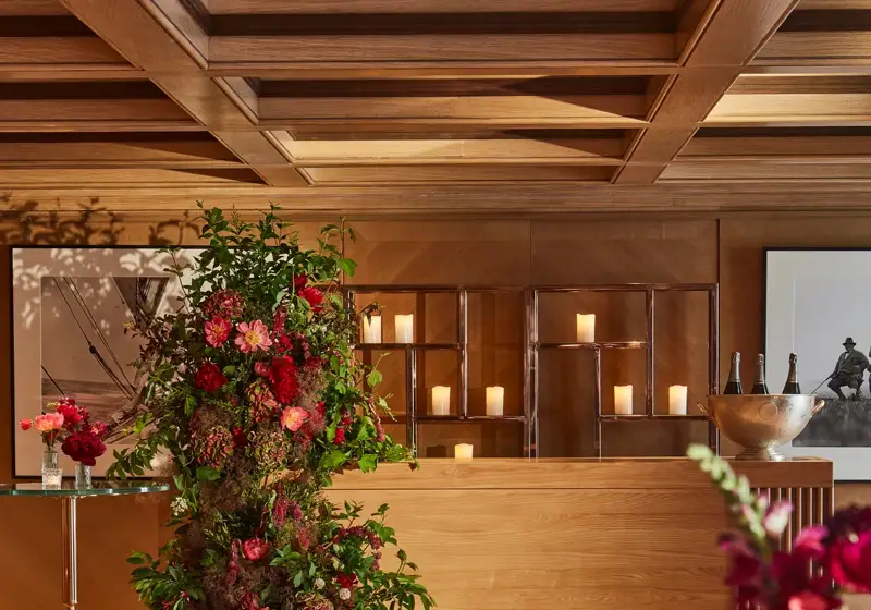 Wood-panelled reception space with a floral installation and candlelit display on a wooden counter, with small tables and flowers in the foreground.