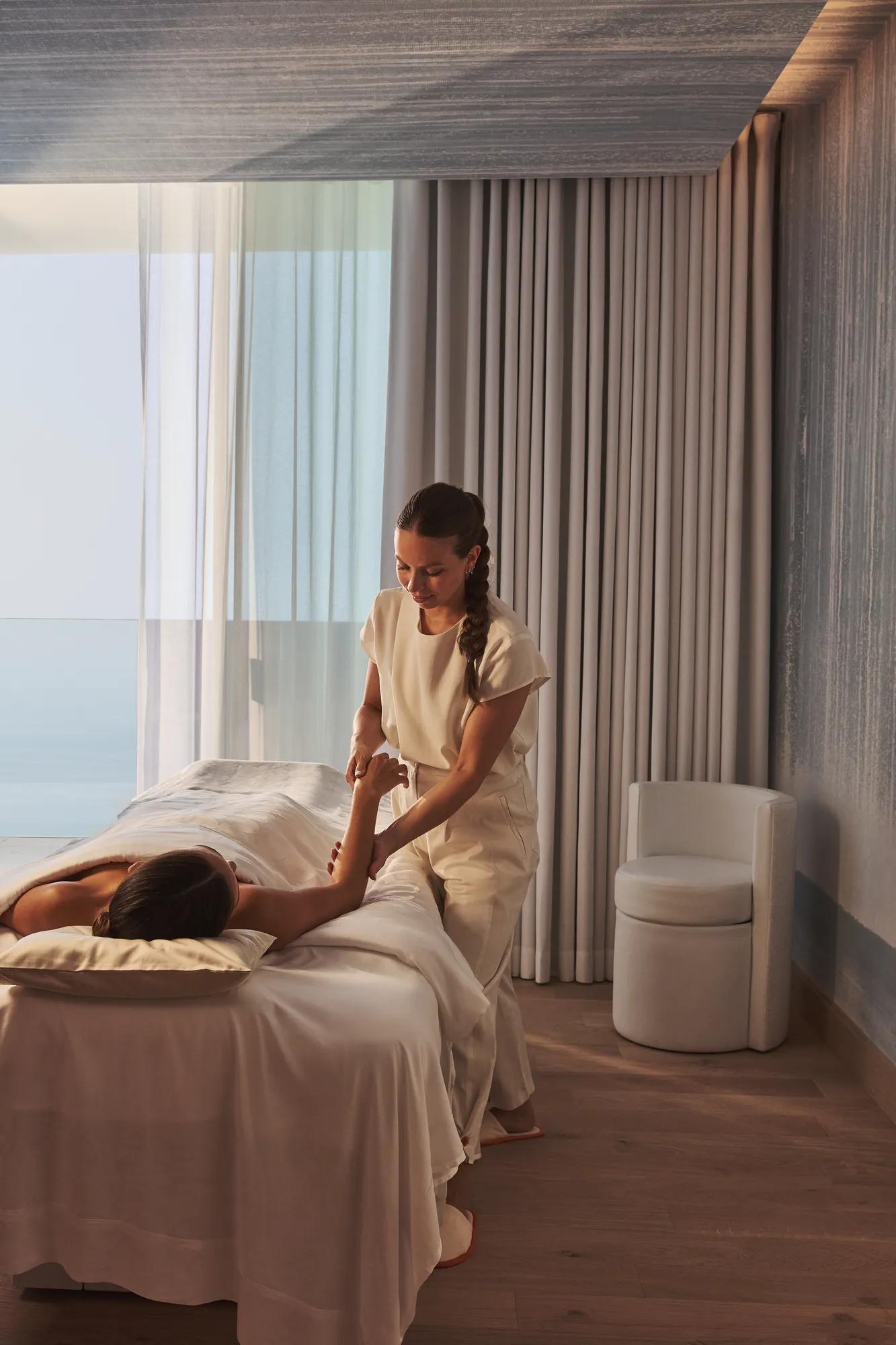 A therapist gives a massage in a serene treatment room with ocean views through floor-to-ceiling windows.
