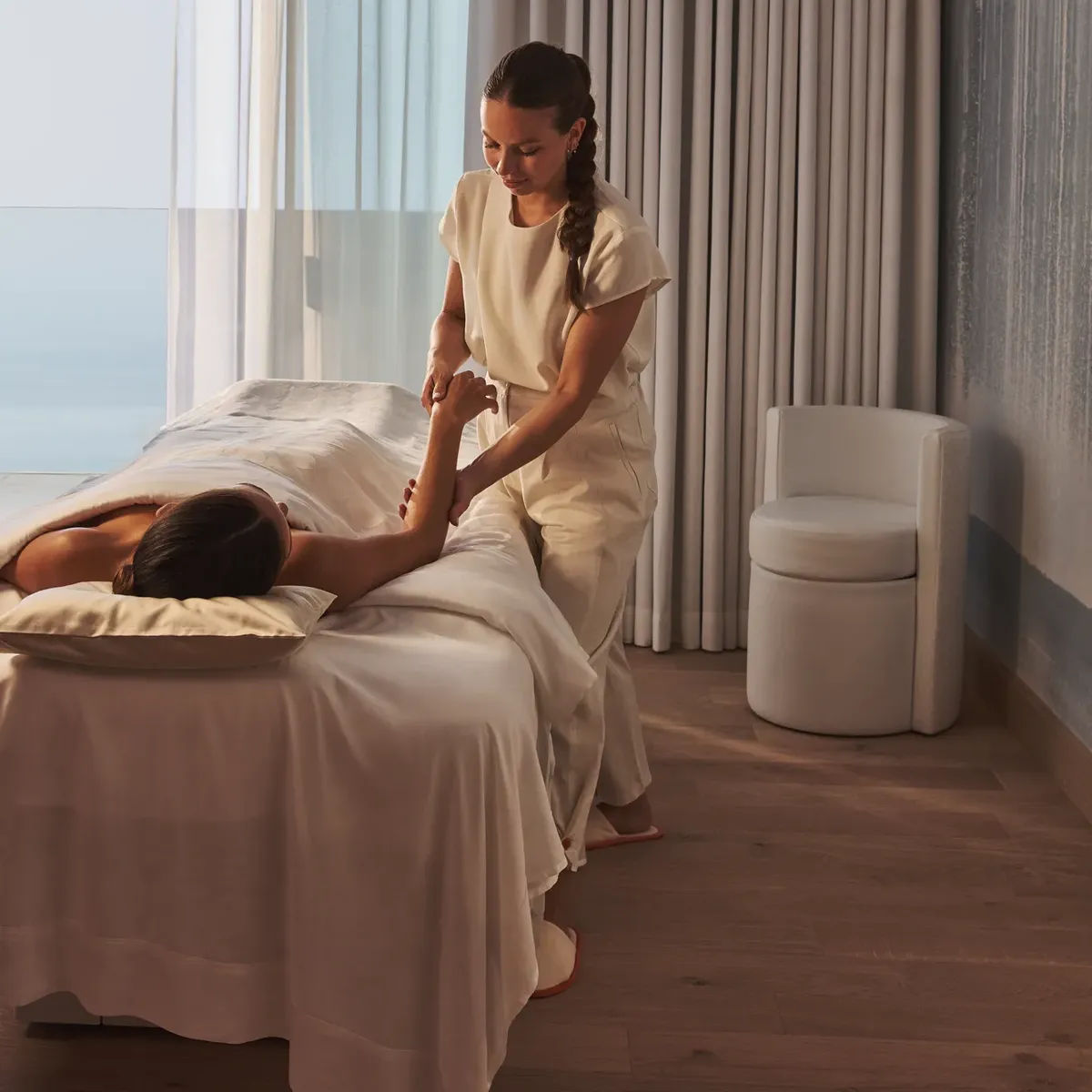A therapist gives a massage in a serene treatment room with ocean views through floor-to-ceiling windows.