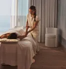 A therapist gives a massage in a serene treatment room with ocean views through floor-to-ceiling windows.