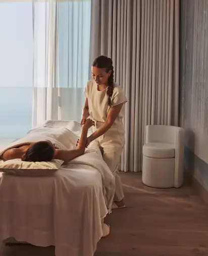 A therapist gives a massage in a serene treatment room with ocean views through floor-to-ceiling windows.