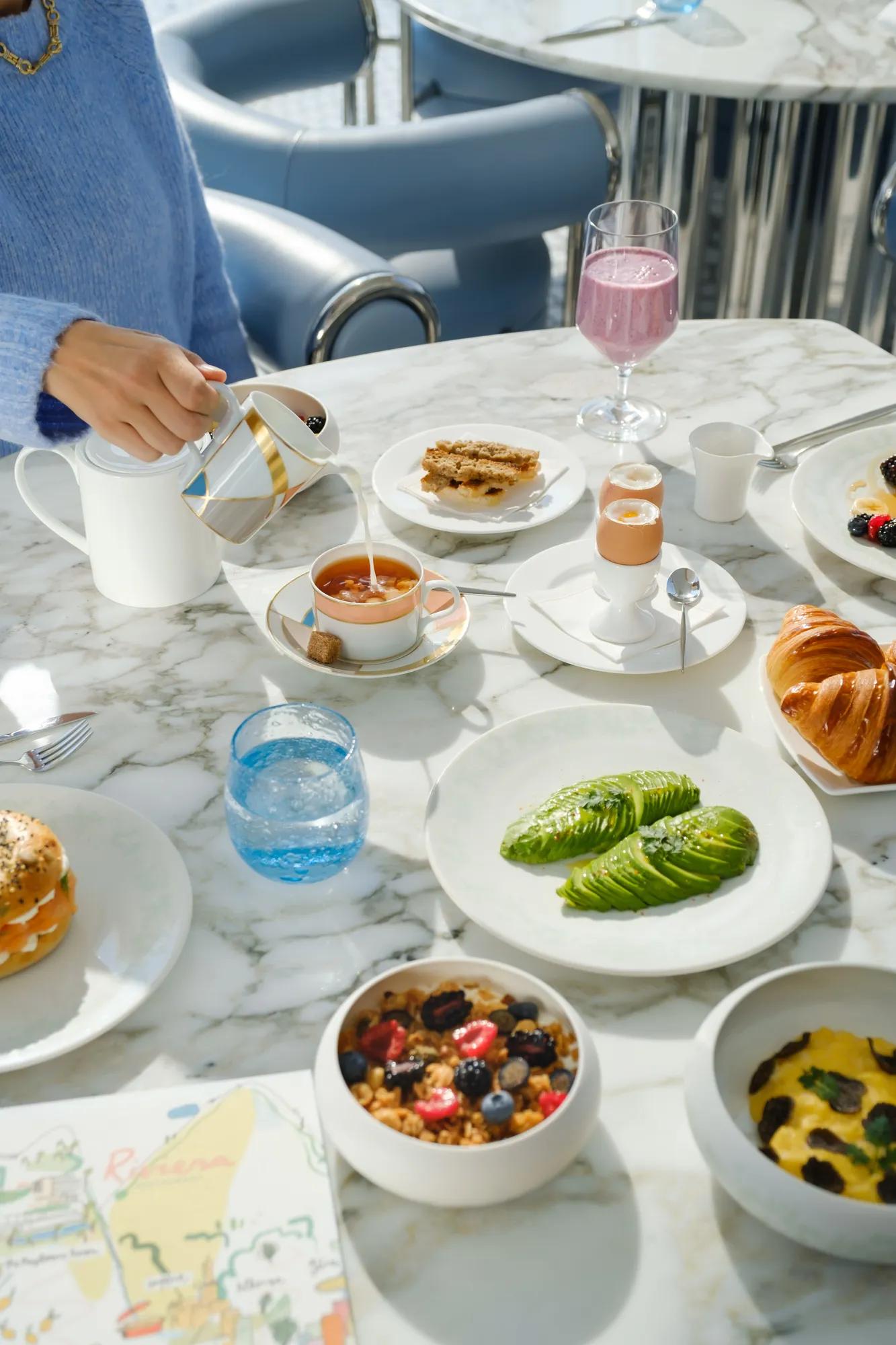 Petit-déjeuner élégant sur table en marbre, avec avocat, pâtisseries, granola, œuf à la coque et main versant du thé.