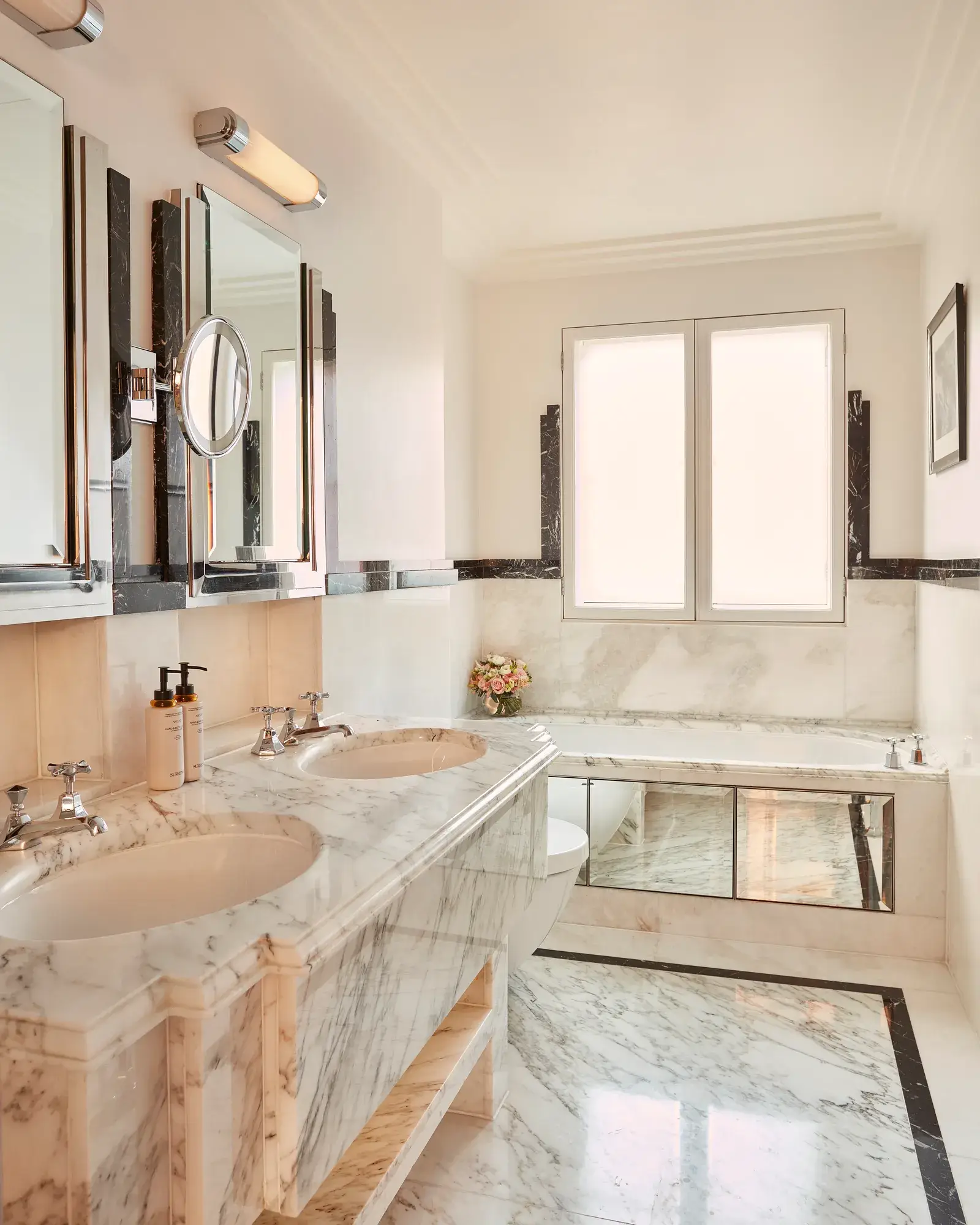 Luxurious marble bathroom with dual sinks, a large bathtub beneath a frosted window, and polished chrome fixtures.