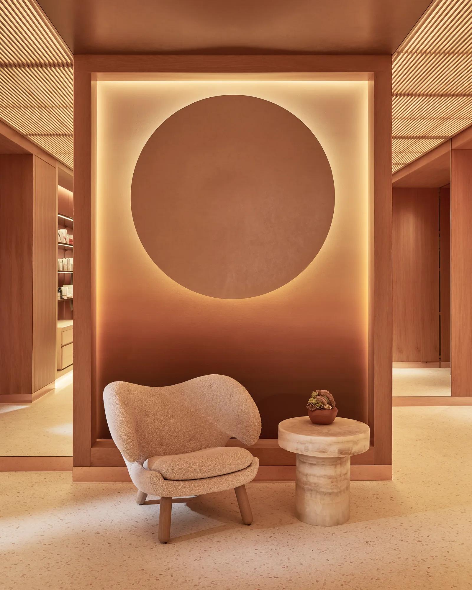 Stylish interior featuring a warm-toned wall with a large illuminated circular design, a plush cream-coloured chair, and a small round table with a potted plant on it, creating a serene and modern atmosphere