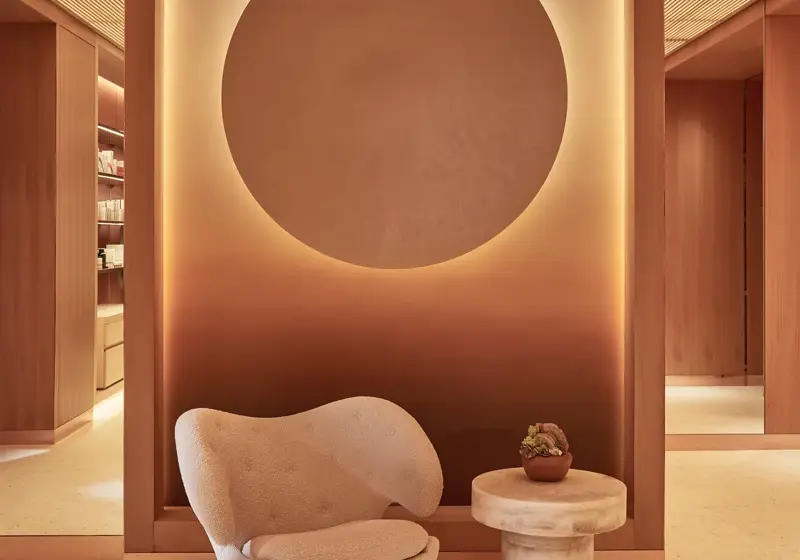 Stylish interior featuring a warm-toned wall with a large illuminated circular design, a plush cream-coloured chair, and a small round table with a potted plant on it, creating a serene and modern atmosphere