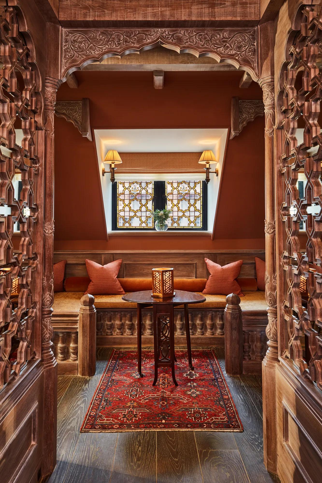 Ornate carved wooden alcove with cushioned seating, lantern table, and patterned red rug.
