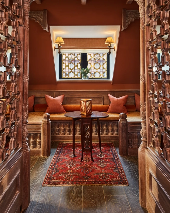 Ornate carved wooden alcove with cushioned seating, lantern table, and patterned red rug.