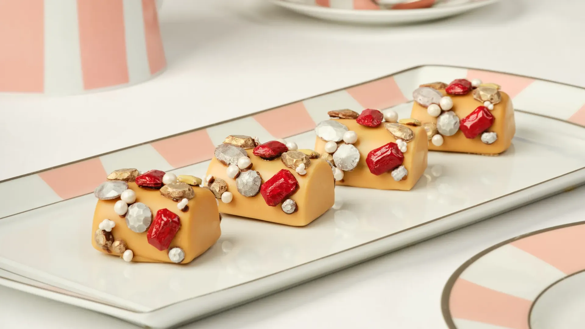 Caramel‑colored pastries topped with red, gold, and silver edible decorations on a striped tray.