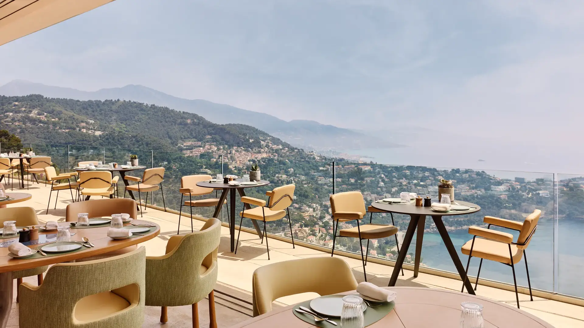 Restaurant en terrasse avec des tables rondes et rectangulaires, des chaises beiges et une vue panoramique sur les collines et le littoral.