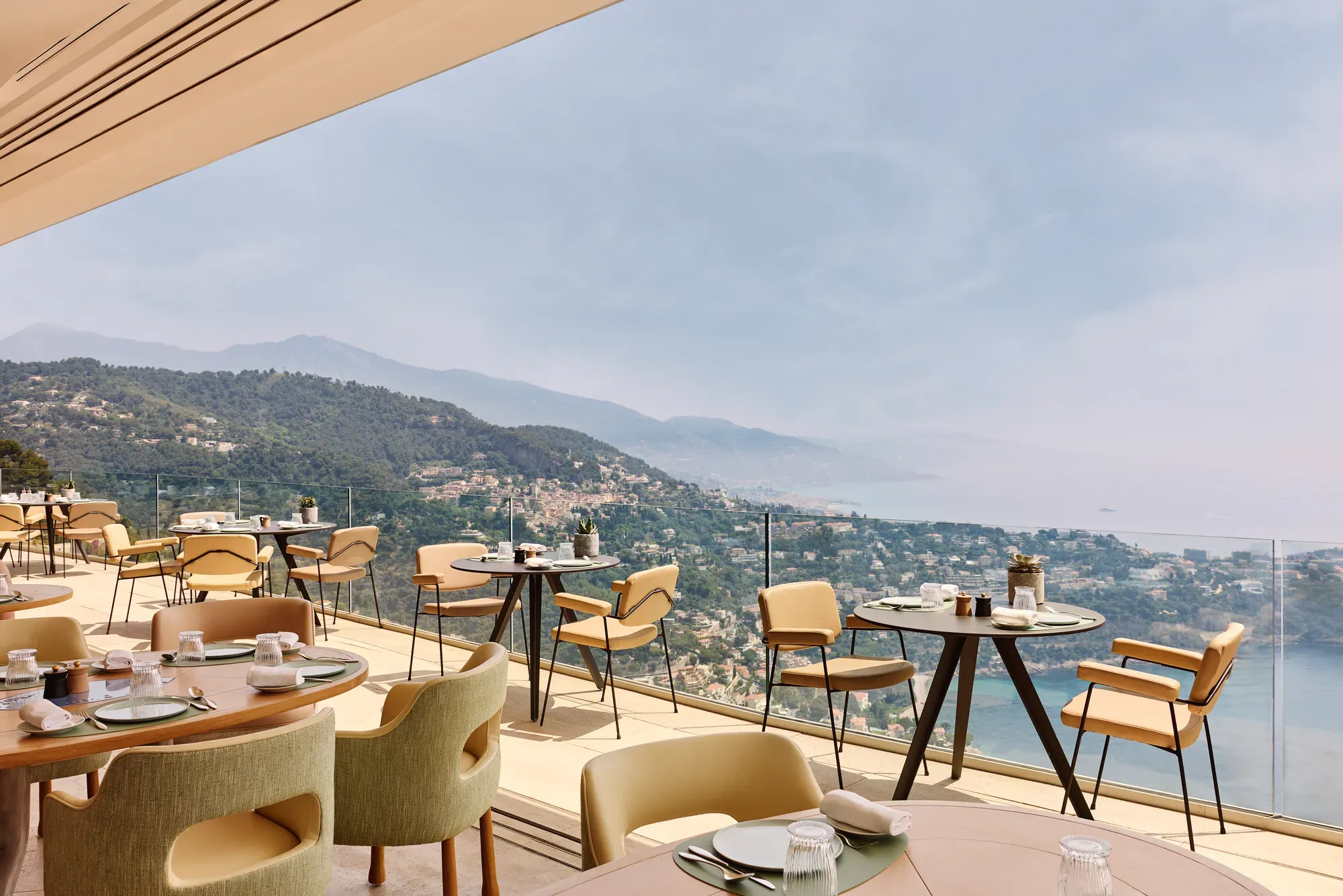 Restaurant en terrasse avec des tables rondes et rectangulaires, des chaises beiges et une vue panoramique sur les collines et le littoral.