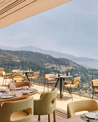 Terrace restaurant with round and rectangular tables, beige chairs, and panoramic views of the hills and coastline.